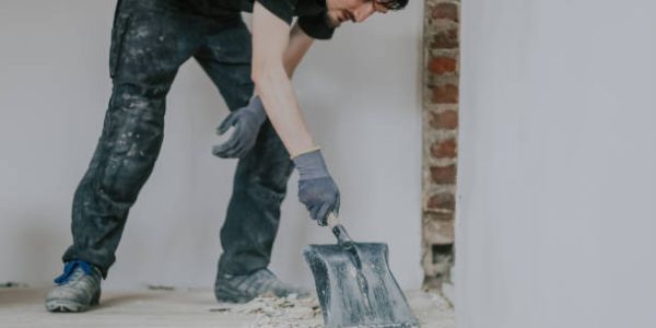 Sloopdiensten One young handsome brunette Caucasian woman in a black uniform stands, bending over, collecting construction debris from the floor with a shovel, close-up side view.